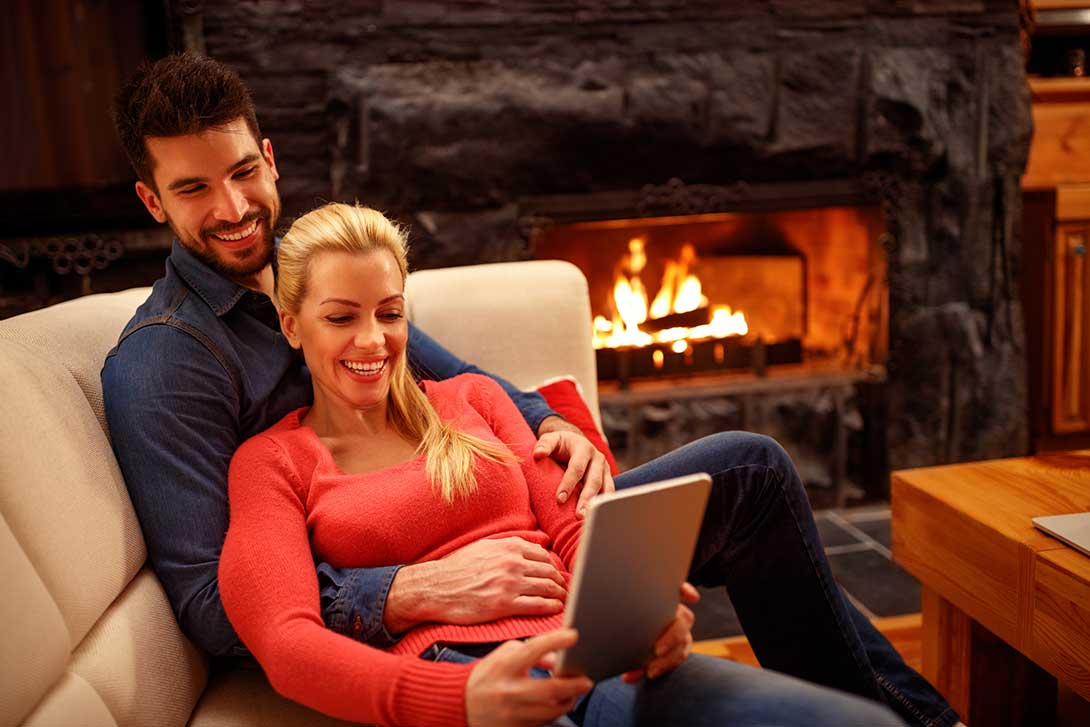 Man and woman sitting on the couch looking at a tablet next to a fireplace